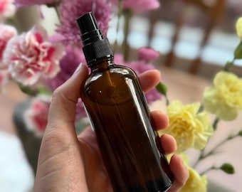 Könnte beinhalten: Eine braune Glassprühflasche mit schwarzem Sprühkopf wird vor einem verschwommenen floralen Hintergrund gehalten. Die Flasche ist mit einer klaren Flüssigkeit gefüllt. Die Blumen sind rosa und gelb.