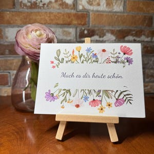 Könnte beinhalten: Eine weiße Karte mit einem floralen Rand und dem Text "Mach es dir heute schön." Die Karte steht auf einem kleinen Holzständer.