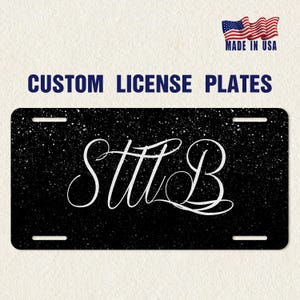 May include: Custom license plate with a black background and white speckled design. The plate features the white cursive letters "STTLB". The words "CUSTOM LICENSE PLATES" are above the plate. The American flag and "MADE IN USA" are in the top right corner.
