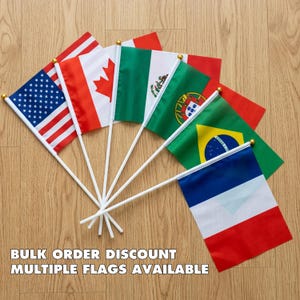 May include: A collection of small flags from various countries, including the United States, Canada, Mexico, Portugal, Brazil, and France. Each flag is mounted on a white stick with a gold-colored top. The flags are arranged on a light brown wooden surface. Text on the image reads: BULK ORDER DISCOUNT MULTIPLE FLAGS AVAILABLE.