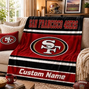 May include: A plush throw blanket featuring the San Francisco 49ers logo in red, black, and white. The design includes the team name in bold red letters at the top, a large central team emblem, and a white text area labeled Custom Name at the bottom.