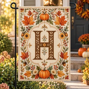 May include: A rectangular garden flag featuring a large, dark brown letter H centered within an ornate frame. The border is decorated with a vibrant autumn pattern of orange maple leaves, pumpkins, sunflowers, acorns, and red berries on a cream background.