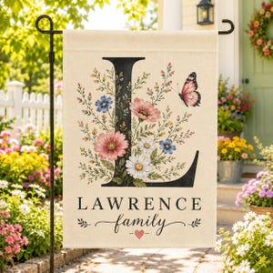 May include: A rectangular garden flag with a cream background featuring a large black letter L decorated with pink, blue, and white wildflowers and a pink butterfly. The text LAWRENCE family is printed below with a small red heart icon.