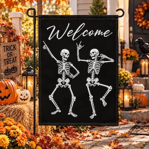 May include: A black rectangular garden flag featuring the white text Welcome at the top and two white skeletons dancing in dynamic poses. The skeletons are depicted with detailed rib cages, skulls, and limbs, set against a solid black background.