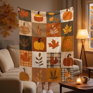 May include: A patchwork throw blanket featuring autumn-themed squares with pumpkins, leaves, and plaid patterns in shades of orange, brown, and green. The blanket is held up, showcasing its design and cozy aesthetic. The blanket is perfect for fall.