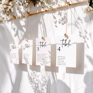 May include: Elegant wedding table number cards. Each card is white with a black script font that reads "Table" followed by a number. The cards are suspended by gold clips and string. The names of guests are listed below the table number.