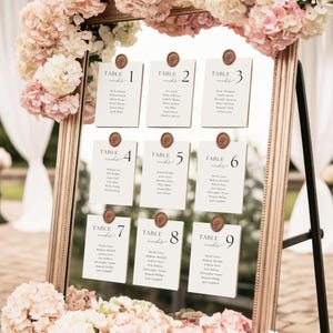 May include: A decorative mirror with a gold frame displays nine table assignment cards for a wedding. Each card is cream-colored with a table number and guest names, sealed with a wax stamp. The mirror is adorned with pink and white flowers.