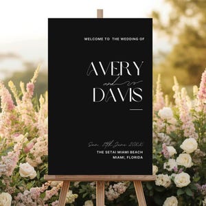 Könnte beinhalten: Schwarzes Hochzeits-Willkommensschild auf einer Holzstaffelei. Der Text in Weiß lautet „WELCOME TO THE WEDDING OF AVERY and DAVIS“ und enthält Veranstaltungsdetails. Das Schild ist von Blumen umgeben.