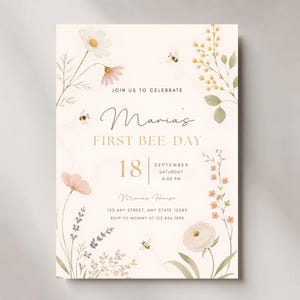 May include: A cream-colored invitation with floral and bee illustrations. The text reads "Maria's FIRST BEE-DAY" with the date, time, and location details. Delicate watercolor flowers and bees decorate the edges, creating a whimsical design.