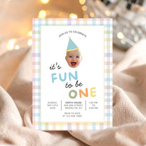 Può includere: Un invito di compleanno dai colori pastello con un bordo a quadri. L'invito presenta un bambino sorridente con un cappello da festa e la scritta "it's FUN to be ONE". Sono inclusi ulteriori dettagli sull'evento.