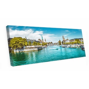May include: A canvas print of a scenic European cityscape. The image features a river with boats, buildings with spires, and a bright blue sky with clouds. The water is a vibrant turquoise color.