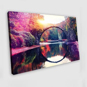 May include: A canvas print depicting a picturesque landscape with a stone bridge spanning a tranquil lake. The scene is filled with the rich colours of autumn, including red, orange, and yellow foliage, reflected in the water.