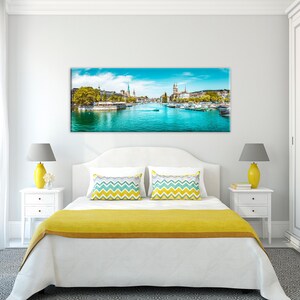 May include: A panoramic canvas print of a European city with a river running through it. The image features a bright blue sky, turquoise water, and buildings with spires. Boats are visible on the water, and trees line the riverbanks.