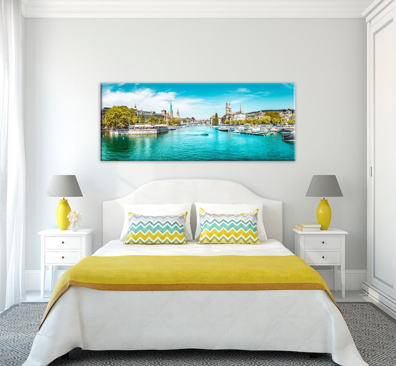 May include: A panoramic canvas print of a European city with a river running through it. The image features a bright blue sky, turquoise water, and buildings with spires. Boats are visible on the water, and trees line the riverbanks.