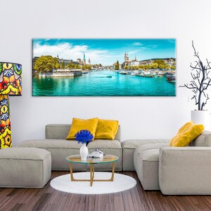 May include: A panoramic canvas print of a cityscape with a bright blue river and sky. The print hangs above a modern gray sectional sofa with yellow pillows. A colorful patterned floor lamp stands to the left.