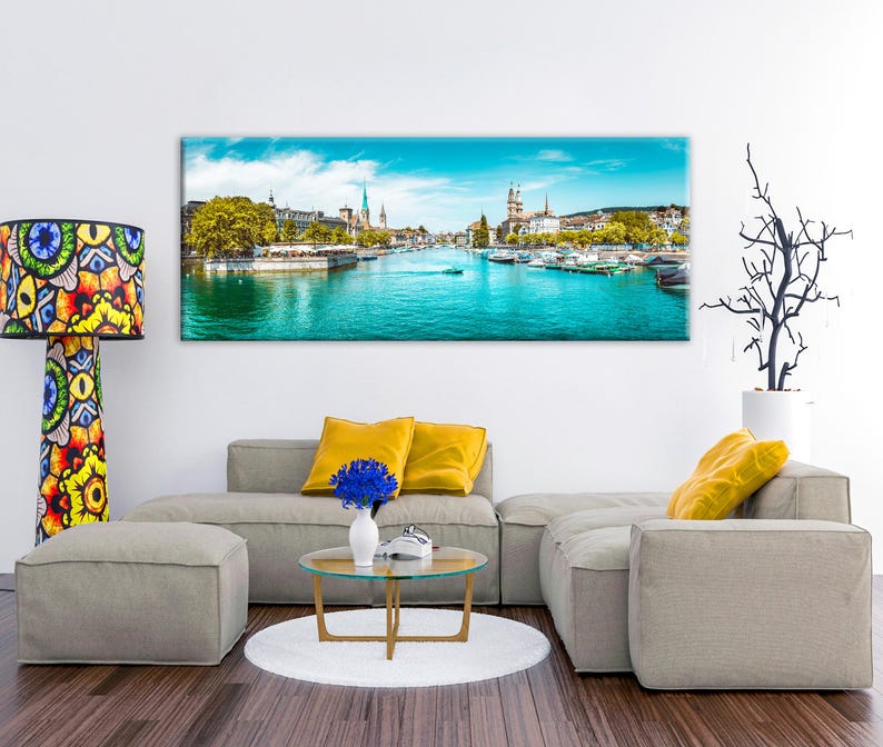 May include: A panoramic canvas print of a cityscape with a bright blue river and sky. The print hangs above a modern gray sectional sofa with yellow pillows. A colorful patterned floor lamp stands to the left.
