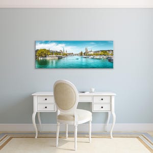 May include: A panoramic canvas print of a scenic European city with a river running through it. The artwork features a bright blue sky, turquoise water, and buildings along the riverbanks. The print is displayed above a white desk and chair.