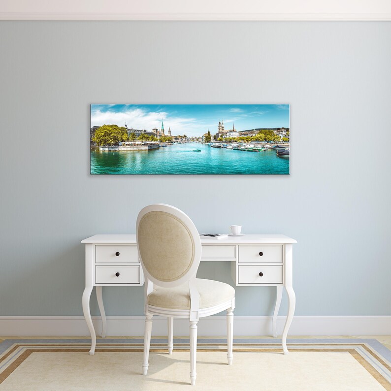 May include: A panoramic canvas print of a scenic European city with a river running through it. The artwork features a bright blue sky, turquoise water, and buildings along the riverbanks. The print is displayed above a white desk and chair.