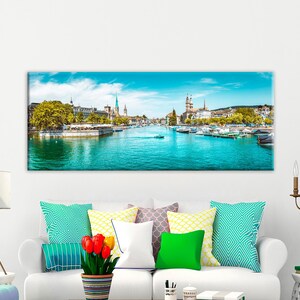May include: A panoramic canvas print of a cityscape featuring a river with boats, buildings, and a bright blue sky. The artwork is displayed above a white sofa with colorful pillows and a lamp.