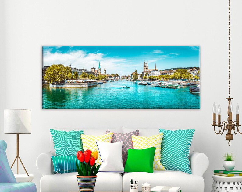 May include: A panoramic canvas print of a cityscape featuring a river with boats, buildings, and a bright blue sky. The artwork is displayed above a white sofa with colorful pillows and a lamp.
