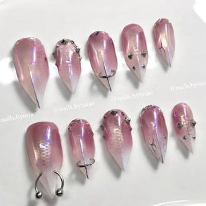 May include: Ten artificial nails with a pink and white gradient design. Some nails have silver studs, rings, or other decorative elements. The text "@nails.bymiao" is visible on the image.