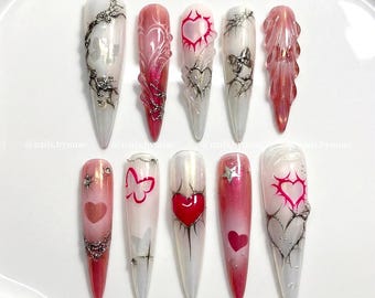 May include: A set of ten long, pointed acrylic nails with various designs. The nails feature a gradient of colors, including red, pink, and white, with heart, butterfly, and abstract patterns. Some nails have silver embellishments.