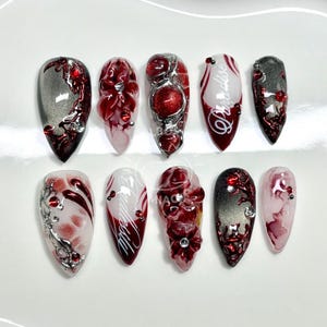 Può includere: Un set di dieci unghie finte con vari design. Le unghie presentano una combinazione di colori rosso, argento e bianco. Alcune unghie hanno accenti floreali, mentre altre mostrano la scritta "Paradise".