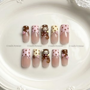 May include: A set of ten press-on nails featuring a French manicure style with a nude base. The tips are decorated with brown, pink, and cream polka dots, and three-dimensional monkey figures. The text @nails.bymiao is visible on the image.