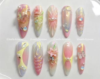 Op de afbeelding: Een verzameling van tien lange, amandelvormige kunstnagels. Elke nagel heeft een uniek ontwerp met iriserende en pastelkleuren, waaronder roze, geel en wit. Versieringen zijn onder meer parels, bloemen en vlinderaccenten.