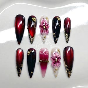 Puede incluir: Un conjunto de diez uñas artificiales largas en forma de almendra. Las uñas presentan un degradado de rojo a negro, detalles dorados y diseños florales. Algunas uñas tienen puntas doradas brillantes y pequeñas cuentas doradas.