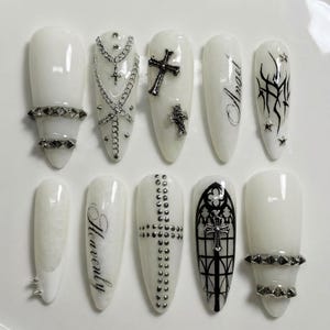 Puede incluir: Un conjunto de diez uñas postizas blancas en forma de almendra con varios diseños. Algunas uñas presentan cadenas, cruces y tachuelas plateadas. Otras tienen las palabras "Angel" y "Heavenly". Una uña tiene un diseño de ventana gótica.