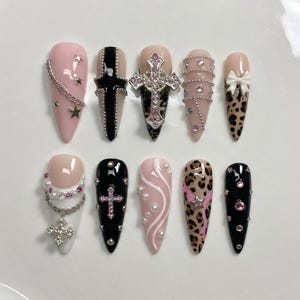 Puede incluir: Un conjunto de diez uñas artificiales largas y puntiagudas. Los diseños incluyen patrones rosas, negros y de leopardo. Los adornos incluyen cadenas plateadas, estrellas, cruces, perlas y pedrería. Las uñas están dispuestas sobre una superficie blanca.