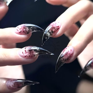 May include: Long, pointed, clear acrylic nails featuring a dark, smoky black gradient tip and a detailed red spider lily floral design painted near the base of each nail. The nails have a sharp, stiletto shape with a glossy finish.