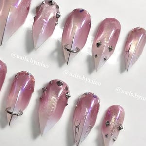 May include: Artificial nails in a pink and white gradient design. Some nails have silver studs, rings, or decorative lines. The image includes the text "@nails.bymiao".