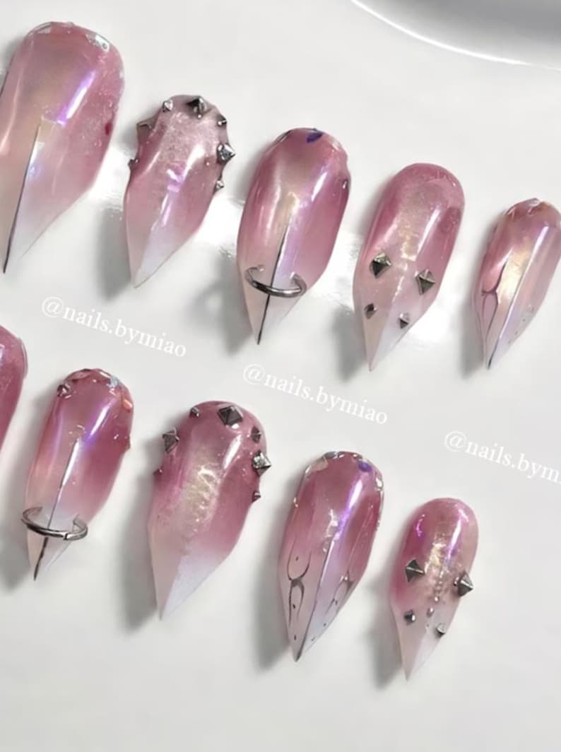 May include: Artificial nails in a pink and white gradient design. Some nails have silver studs, rings, or decorative lines. The image includes the text "@nails.bymiao".