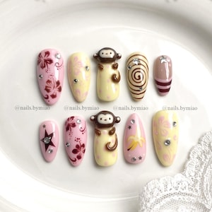 May include: A set of ten almond-shaped press-on nails featuring various designs including pink hibiscus flowers, brown monkey figures, yellow bananas, brown swirls, and stars. The nails are decorated with small clear rhinestones and the text @nails.bymiao.