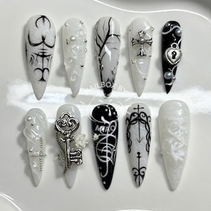 Puede incluir: Un conjunto de diez uñas artificiales largas y puntiagudas. Los diseños incluyen arte gótico en blanco y negro, cruces, llaves y patrones abstractos. Algunas uñas presentan adornos 3D como perlas y dijes. Las uñas están dispuestas sobre una superficie blanca.