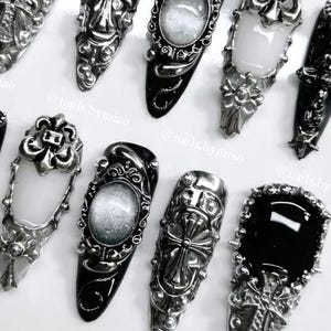 Op de afbeelding: Een assortiment zwarte en zilveren nail art-ontwerpen. De nagels zijn voorzien van sierlijke zilveren versieringen, waaronder kruisen, bloempatronen en ovale frames. De nagellak is glanzend zwart, met sommige nagels met een wit of zilver interieur.