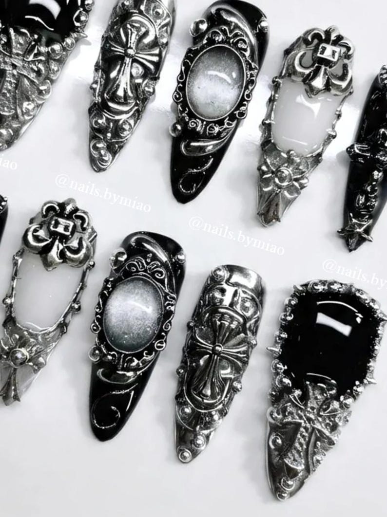 Op de afbeelding: Een assortiment zwarte en zilveren nail art-ontwerpen. De nagels zijn voorzien van sierlijke zilveren versieringen, waaronder kruisen, bloempatronen en ovale frames. De nagellak is glanzend zwart, met sommige nagels met een wit of zilver interieur.