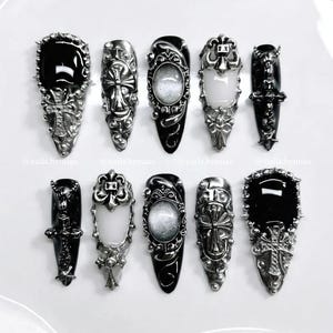 Op de afbeelding: Collectie van tien gothic-stijl nail art ontwerpen. Elke nagel heeft een zwarte basis met zilveren versieringen, waaronder kruisen en ovale accenten. De nagels zijn gerangschikt op een wit oppervlak. De ontwerpen zijn gedetailleerd.