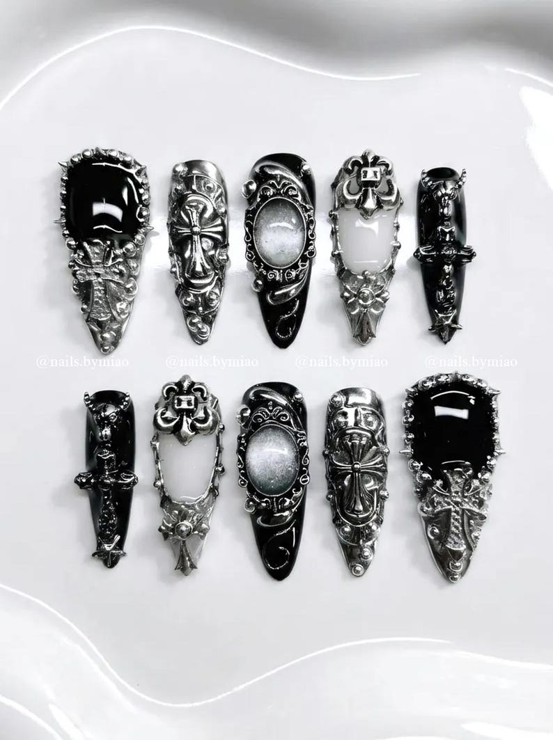 Op de afbeelding: Collectie van tien gothic-stijl nail art ontwerpen. Elke nagel heeft een zwarte basis met zilveren versieringen, waaronder kruisen en ovale accenten. De nagels zijn gerangschikt op een wit oppervlak. De ontwerpen zijn gedetailleerd.