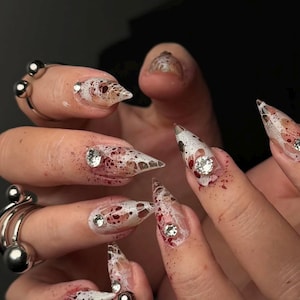 Peut inclure: Gros plan sur des ongles en acrylique longs et pointus avec un design unique. Les ongles présentent une base transparente avec des motifs blancs en forme de dentelle, des mouchetures rouges et des strass étincelants. Des bagues argentées ornent les doigts.