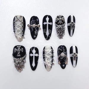 May include: A set of ten black and white press-on nails with various designs. Some nails feature crosses, lace-up details, and silver embellishments. The nails are oval-shaped and have a glossy finish, showcasing a gothic aesthetic.