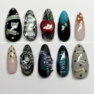 May include: A collection of ten almond-shaped nail designs. The nails feature various designs, including text, polka dots, an apple, and floral accents. Colors include black, white, red, blue, and neutral tones. The text on the nails reads "so the lion fell in love", "twilight", and "with the lamb".