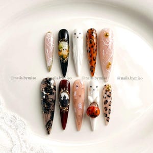 May include: A set of ten stiletto-shaped press-on nails featuring diverse designs including white lace patterns, a black nail with a ghost cameo, a white ghost, leopard print, a black lace overlay, a dark red nail with a haunted house cameo, and a ghost holding a pumpkin bucket. The text @nails.bymiao is visible.