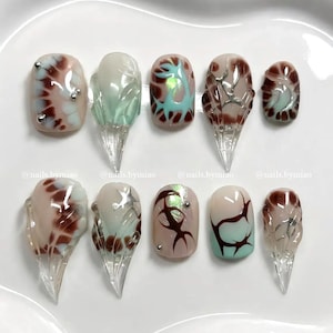 May include: A collection of ten artificial nails with various designs. The nails feature a combination of colors, including brown, beige, and teal, with silver accents. Some nails have a clear, pointed extension. The text "@nails.bymiao" is visible.