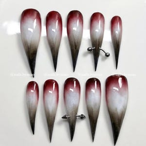 Può includere: Un set di dieci unghie finte lunghe a forma di mandorla. Ogni unghia presenta un design sfumato, che passa da un rosso intenso alla cuticola al bianco al centro, sfumando in un marrone scuro sulla punta. Alcune unghie hanno hardware argentato.