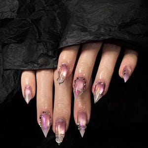 May include: Close-up of a hand with long, pointed, clear acrylic nails. The nails have a gradient effect, transitioning from pink to clear. Some nails are adorned with silver studs and a silver ring.