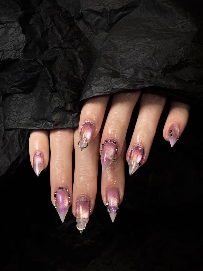 May include: Close-up of a hand with long, pointed, clear acrylic nails. The nails have a gradient effect, transitioning from pink to clear. Some nails are adorned with silver studs and a silver ring.