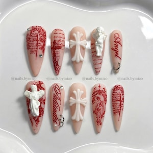 May include: A set of ten long, almond-shaped artificial nails. The nails feature a pale pink base with various designs, including red script, crosses, angel figurines, and the word "Angel". Some nails have silver rings or rhinestones.